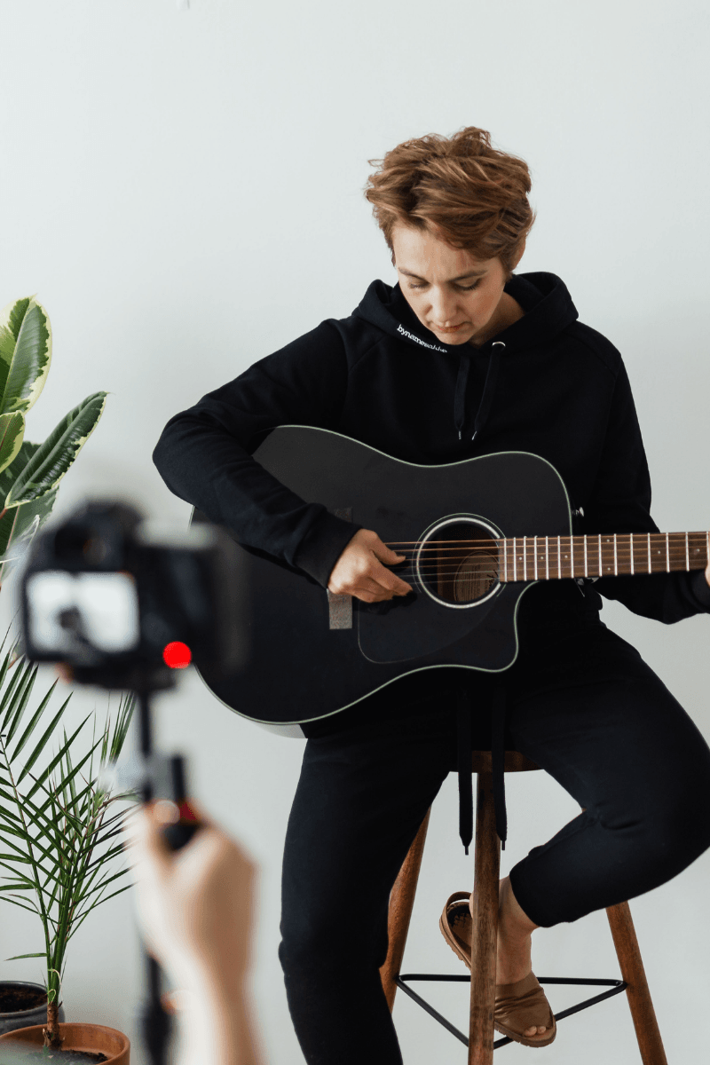 Guitarist recording a video performance while a camera films in a minimal indoor studio space.
