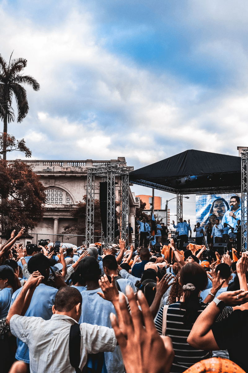 Large outdoor crowd with raised hands facing a stage at a daytime music event.